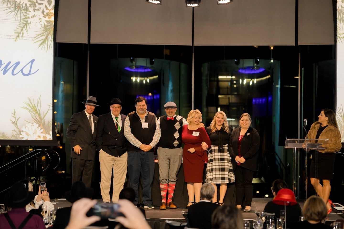 Group of people on stage, some with festive attire. Woman speaking at a podium, others smiling. Interior setting.