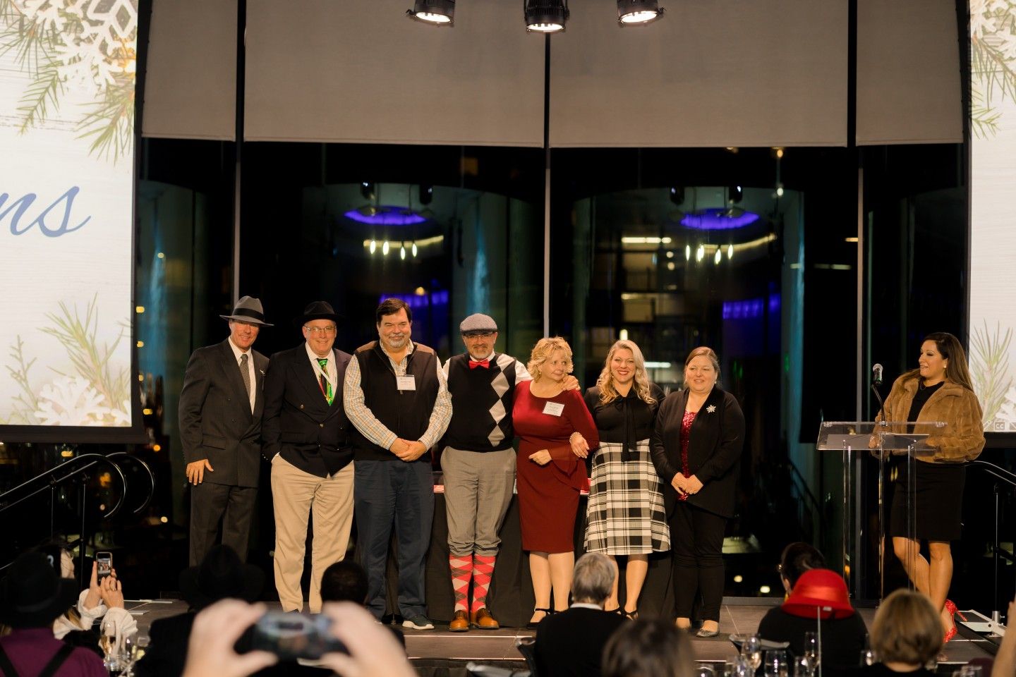 Group of people on stage, dressed up. Some are holding awards. Background has festive decor.