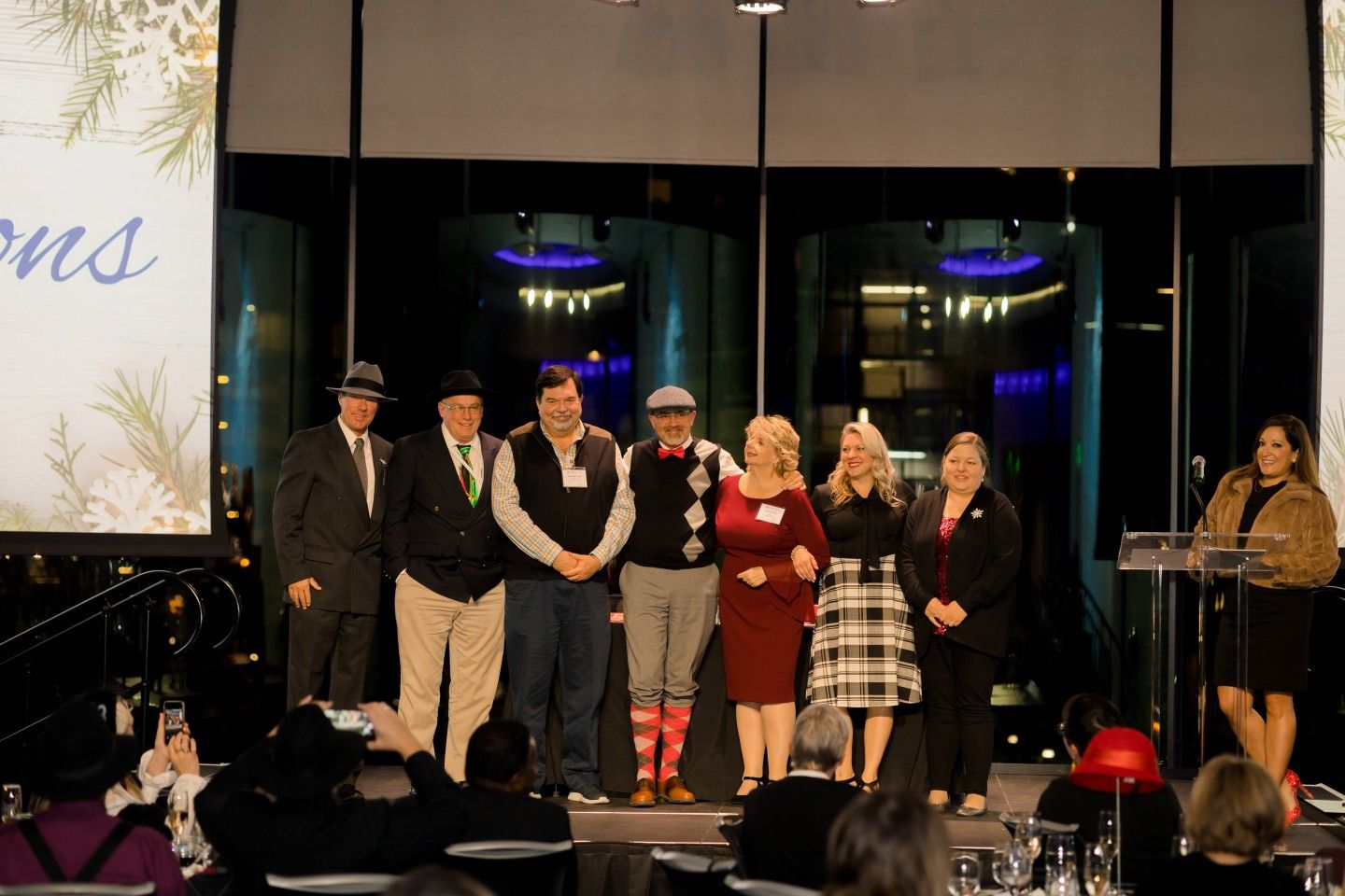 Group of people on stage at an event, some wearing hats. A woman speaks at a podium.