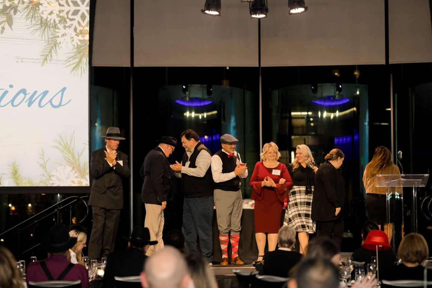 People on a stage at an event, accepting awards.  Guests seated at tables in a brightly lit room.