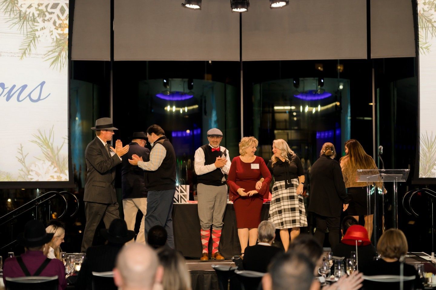 People on stage at an awards event, clapping. Guests in formal attire. Stage decorations include floral design and branding.