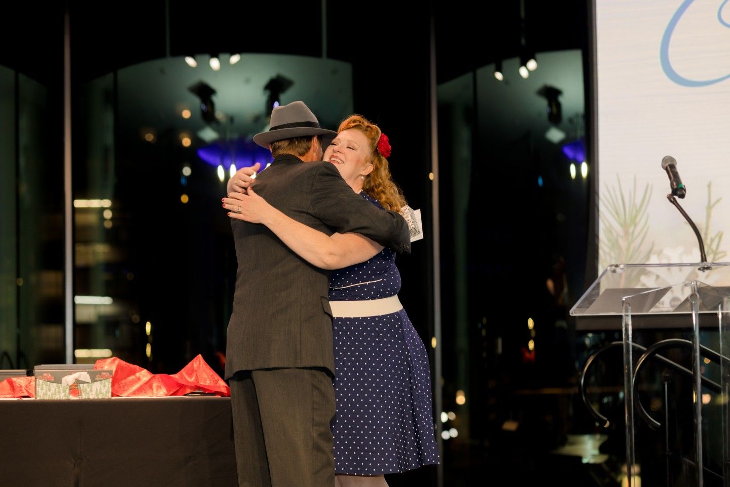 Two people hugging onstage; woman in polka dot dress, man in suit and hat.