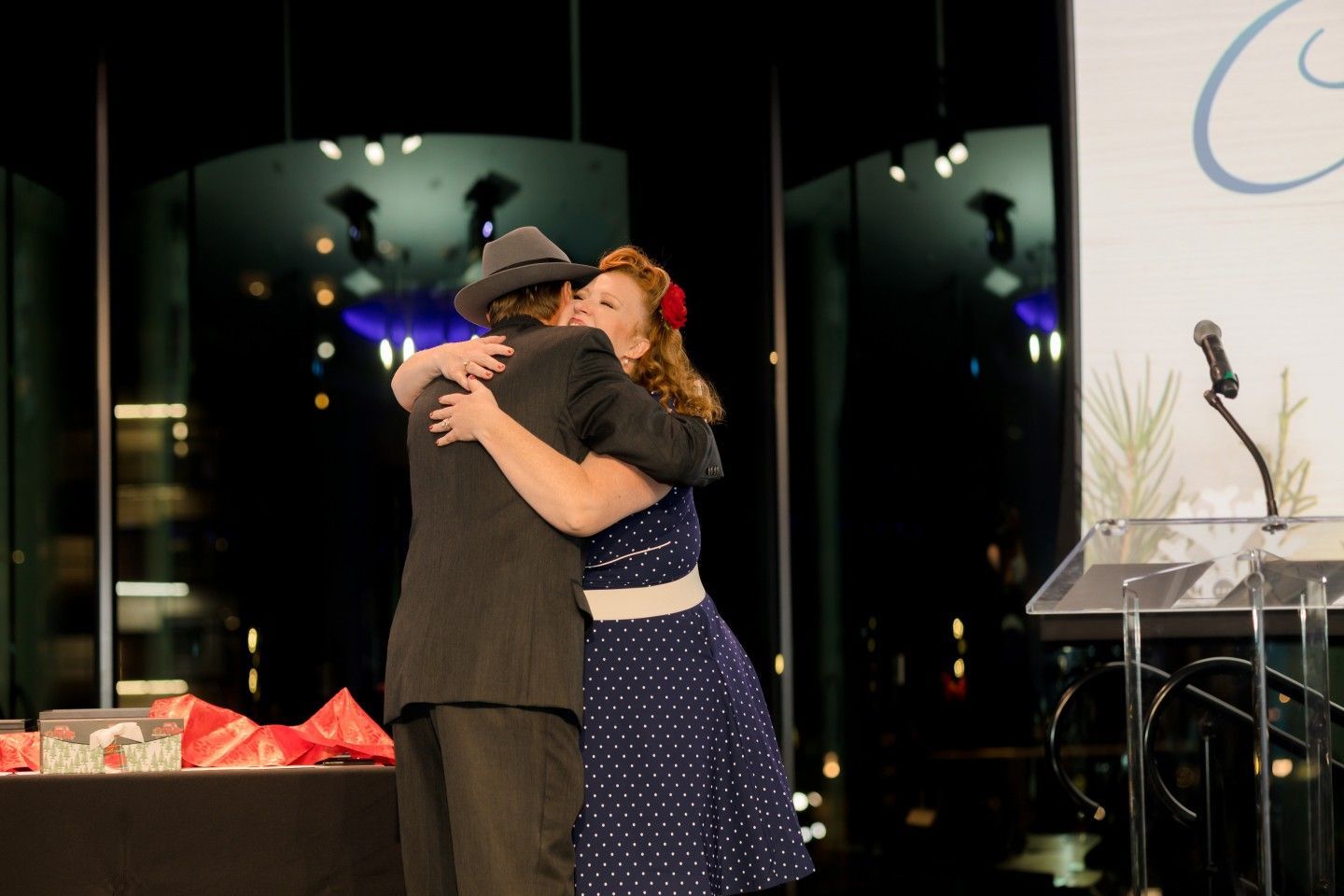Man in fedora hugs woman in polka dot dress on stage.