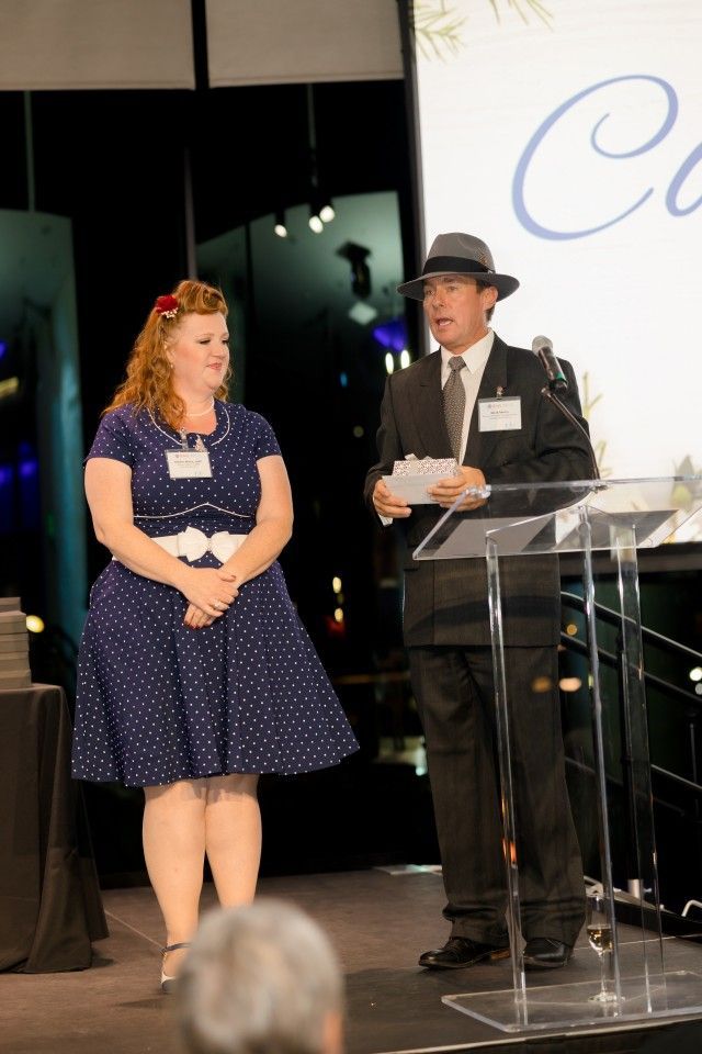 Woman in polka dot dress and man in suit stand at podium; they appear to be at an event.