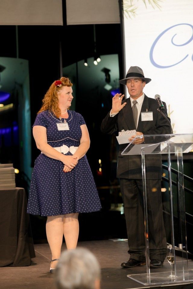 Two people on stage at an event. Woman in blue polka dot dress; man in suit and fedora gesturing.