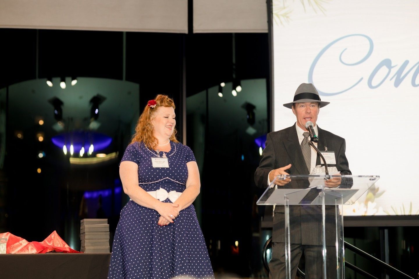 Man in fedora speaks at podium, woman in polka dots stands beside him at an event.
