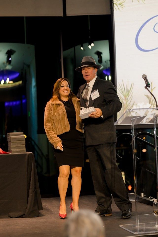 Woman in black dress and fur coat with man in fedora holding award on stage.