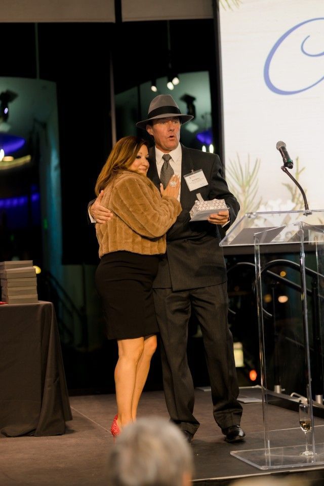 Woman hugging a man on stage; he holds award and wears a fedora. They're near a podium.