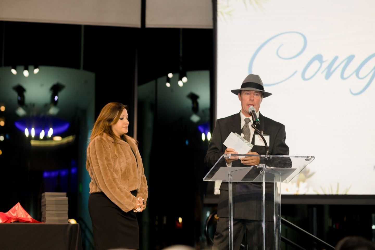 Man in suit and fedora speaks at podium, woman stands beside him at event with 