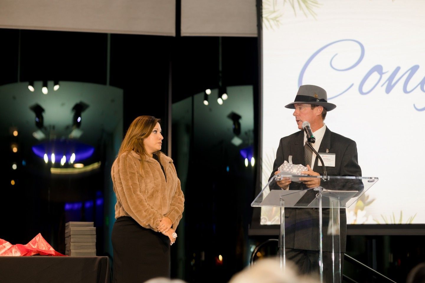 Man in suit with hat speaks at podium, presenting award to woman in fur coat.