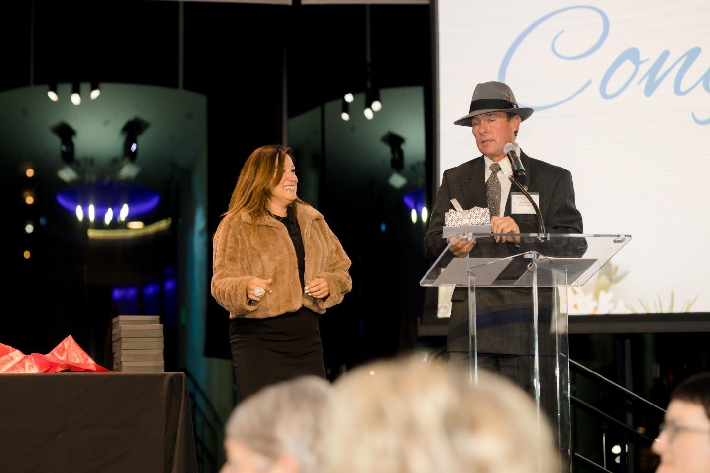 Man in fedora speaking at podium, woman smiling nearby at event.
