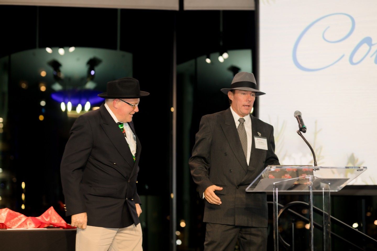 Two men in suits and hats at a podium, one speaking. Nighttime event with a lit background.