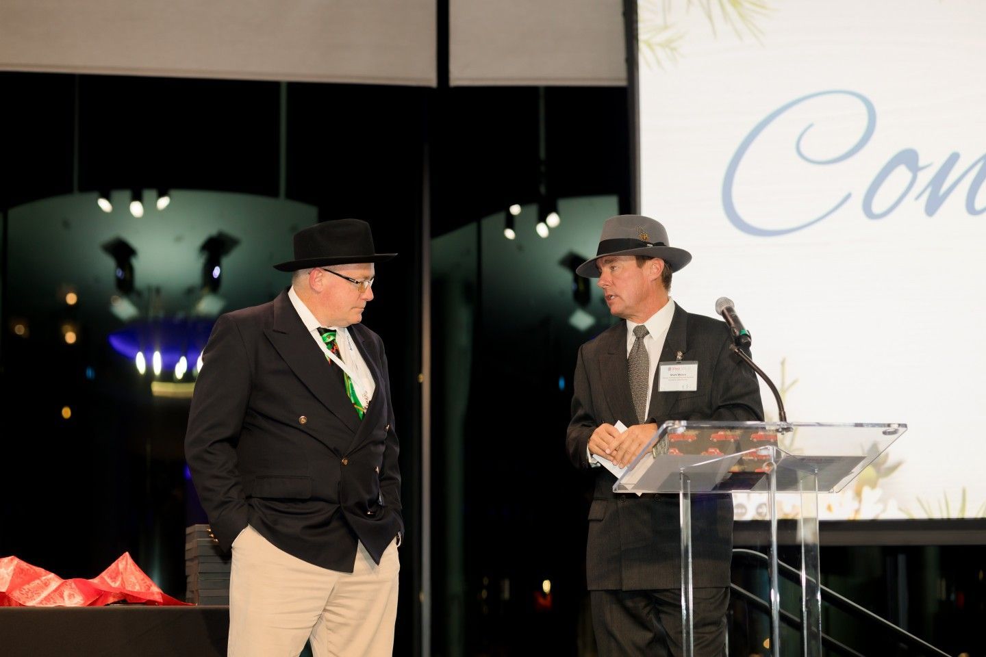 Two men in hats at a podium. One speaks into a microphone. Dark suits, backdrop says 