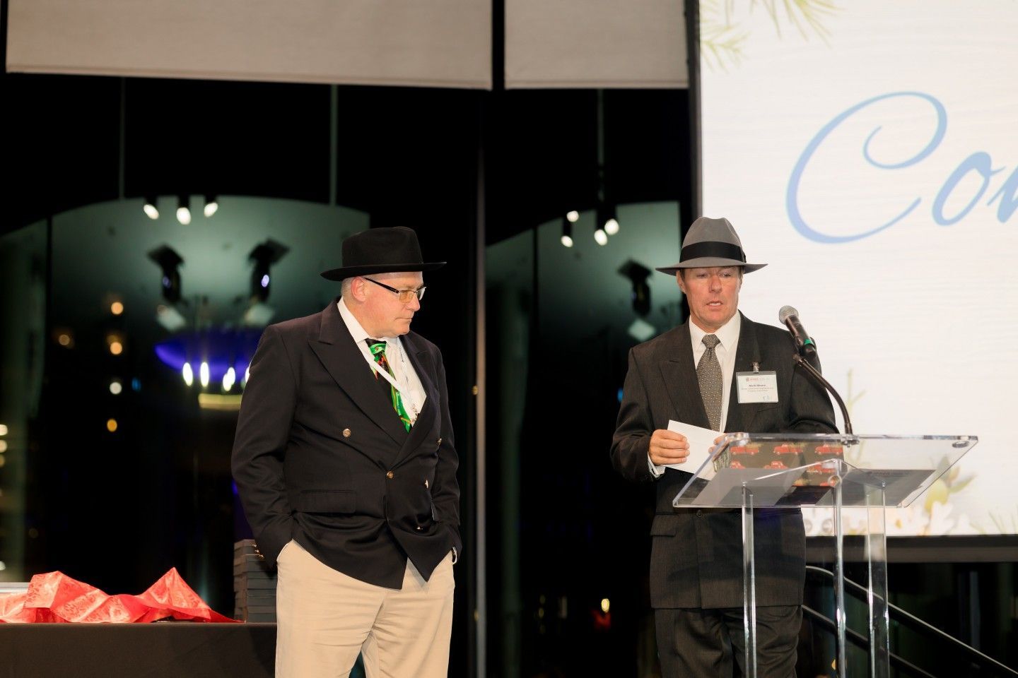 Two men in hats at a podium. One speaks, the other stands beside him.  Indoor setting with a screen.