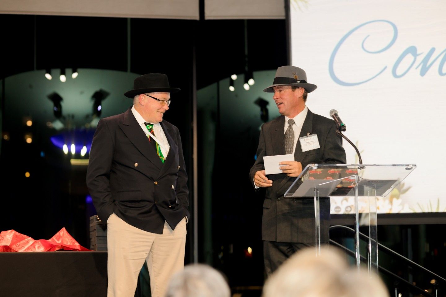 Two men in hats at a podium, one speaking. Nighttime event with a lit backdrop and a 