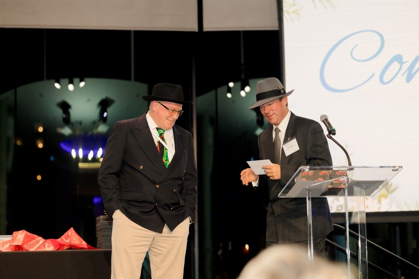 Two men in hats at a podium. One holds papers, the other smiles. Stage, white background, night setting.
