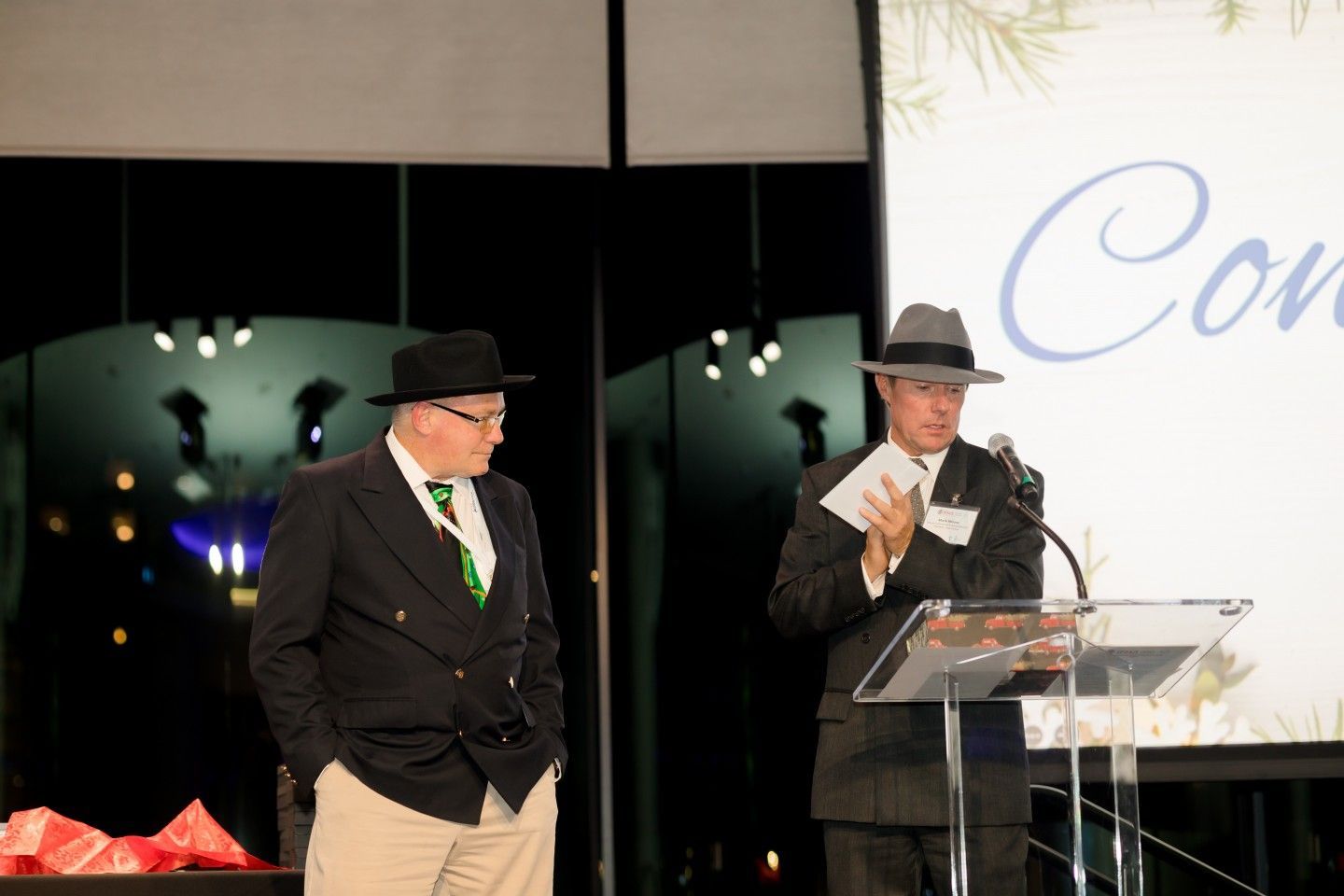 Two men in hats at a podium. One speaks into a microphone, the other stands beside him, both in suits.