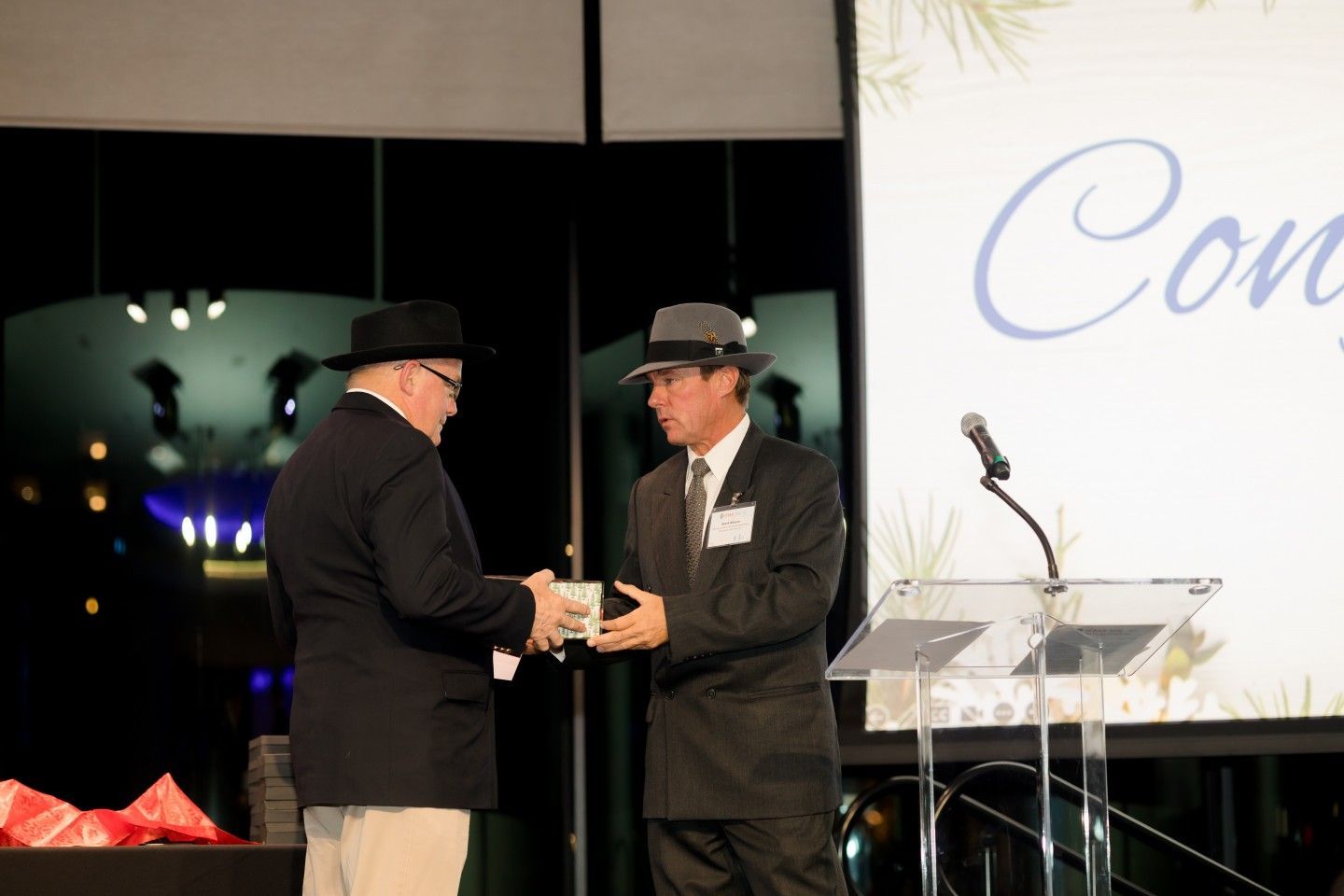 Two men in suits and hats exchange a gift box on a stage at an event.