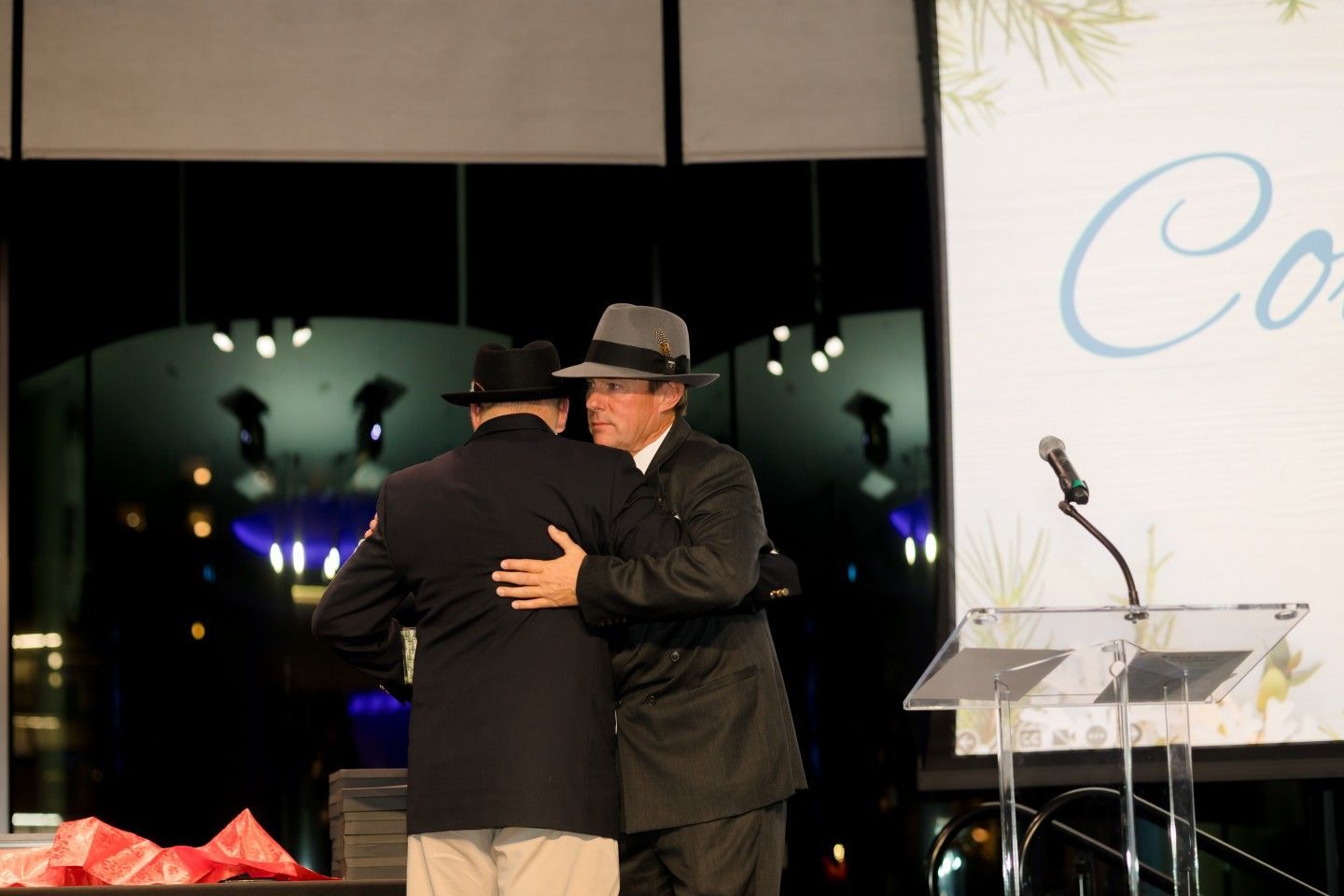 Two men in fedora hats embrace on a stage; a microphone and backdrop are visible.