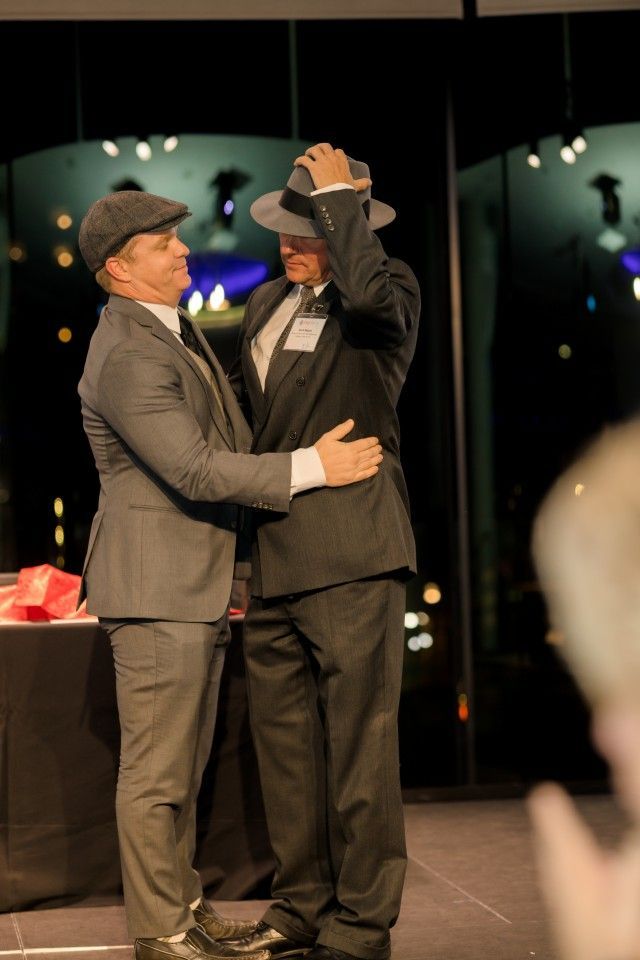 Two men in suits embrace onstage, exchanging hats, celebratory event.