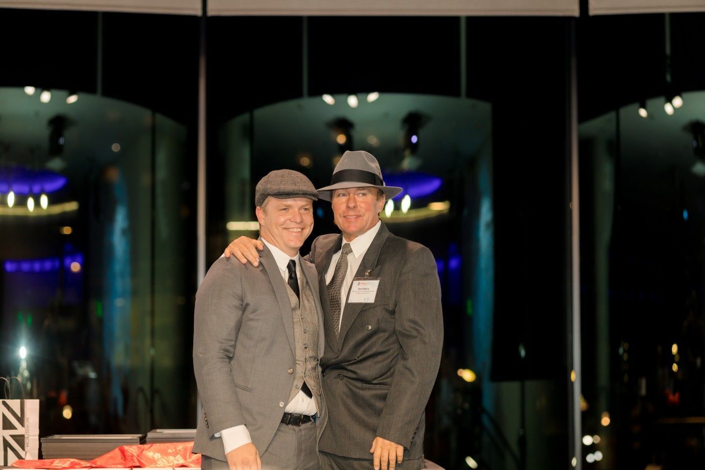 Two men in suits and hats pose together, one with arm around the other at an event.