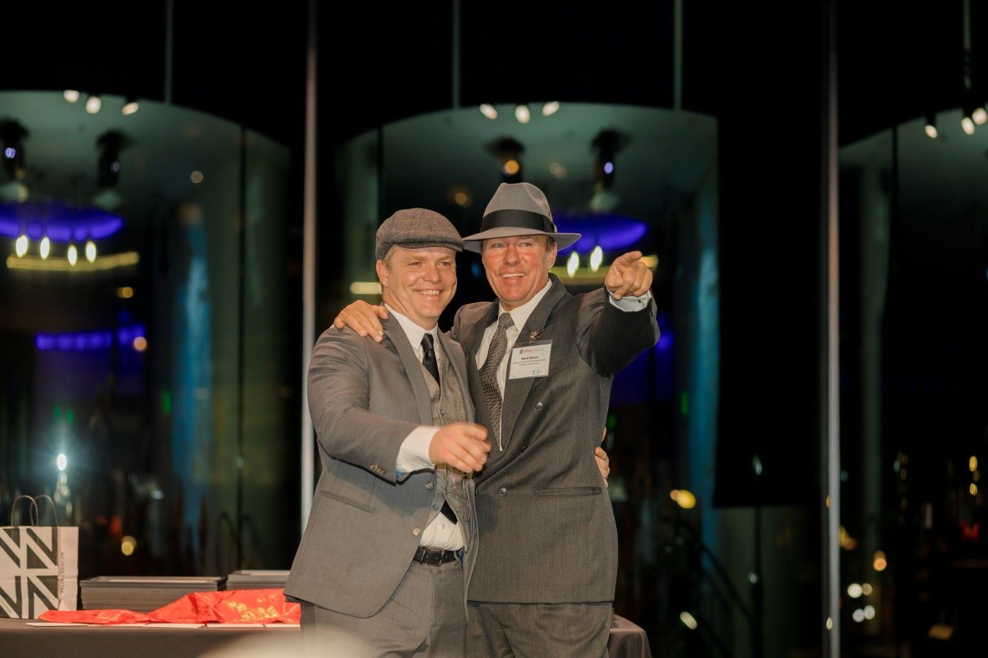 Two men in suits and hats on a stage, one pointing, with city lights visible in background.