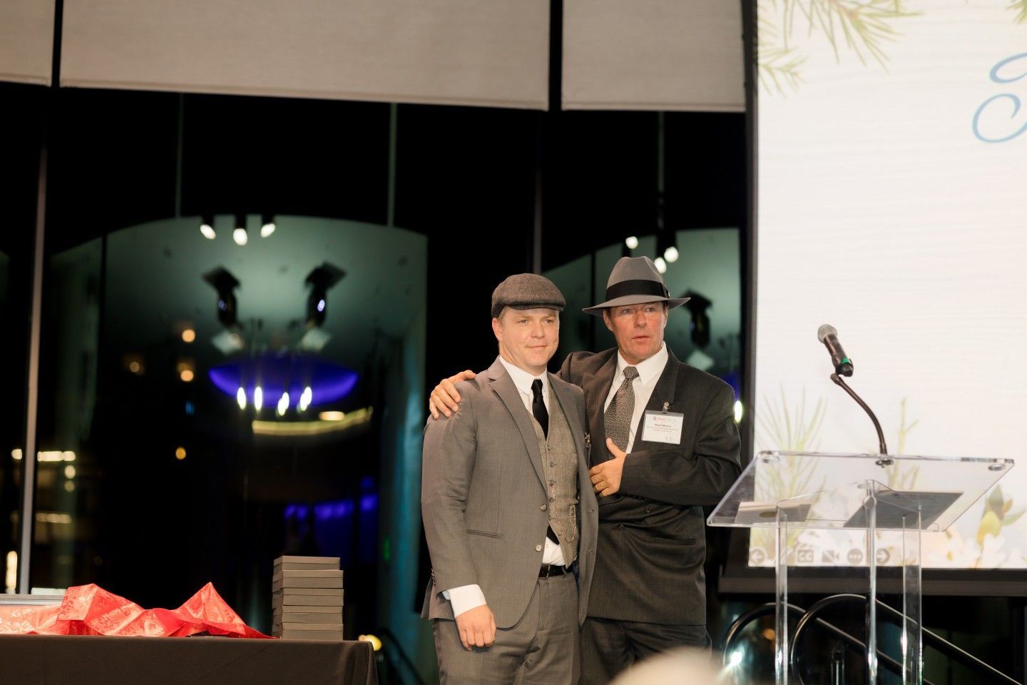 Two men in suits and hats on stage at an event, one with an arm around the other.