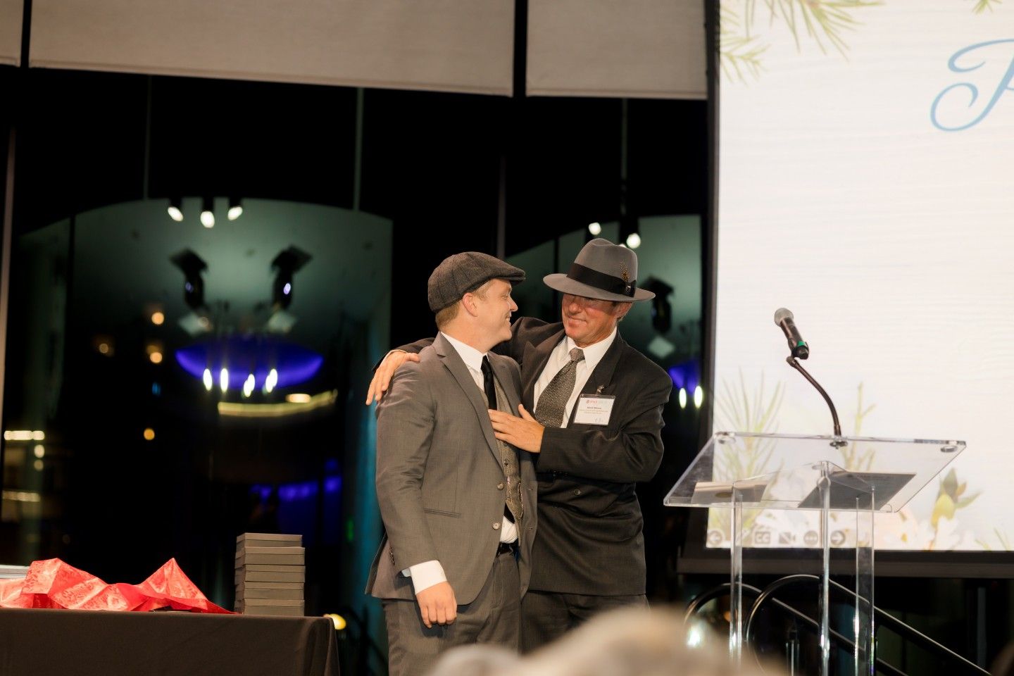 Two men in suits and hats on stage, one with arm around the other; microphone and podium.