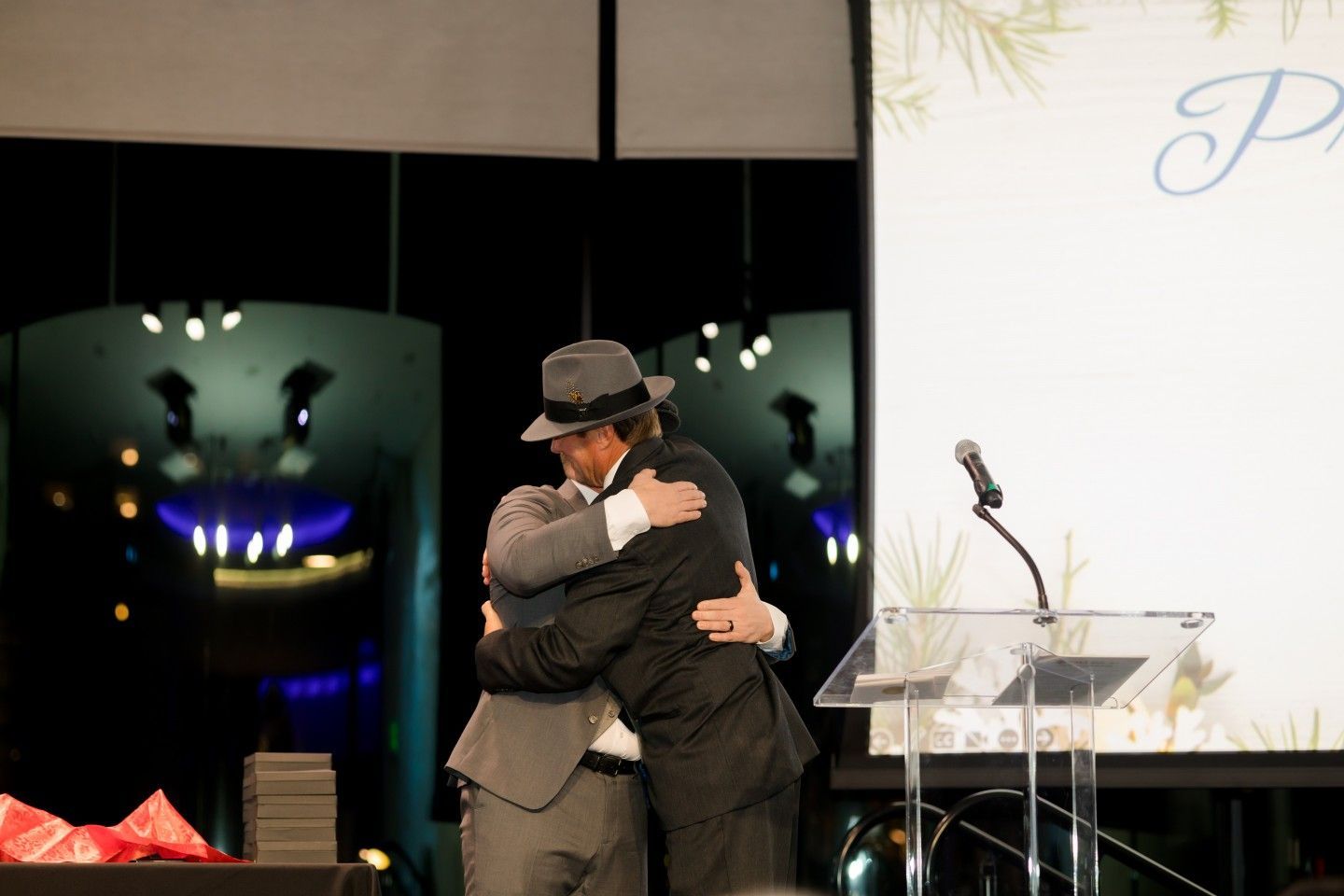Two men embrace on stage at an event. One wears a fedora. A podium and screen are visible.