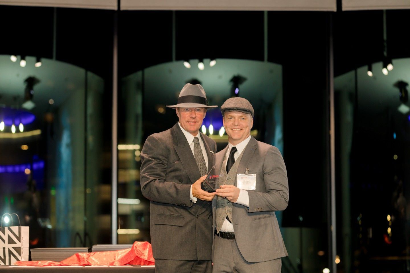 Two men in suits and hats on stage, one holding an award, smiling. Nighttime setting with city lights.