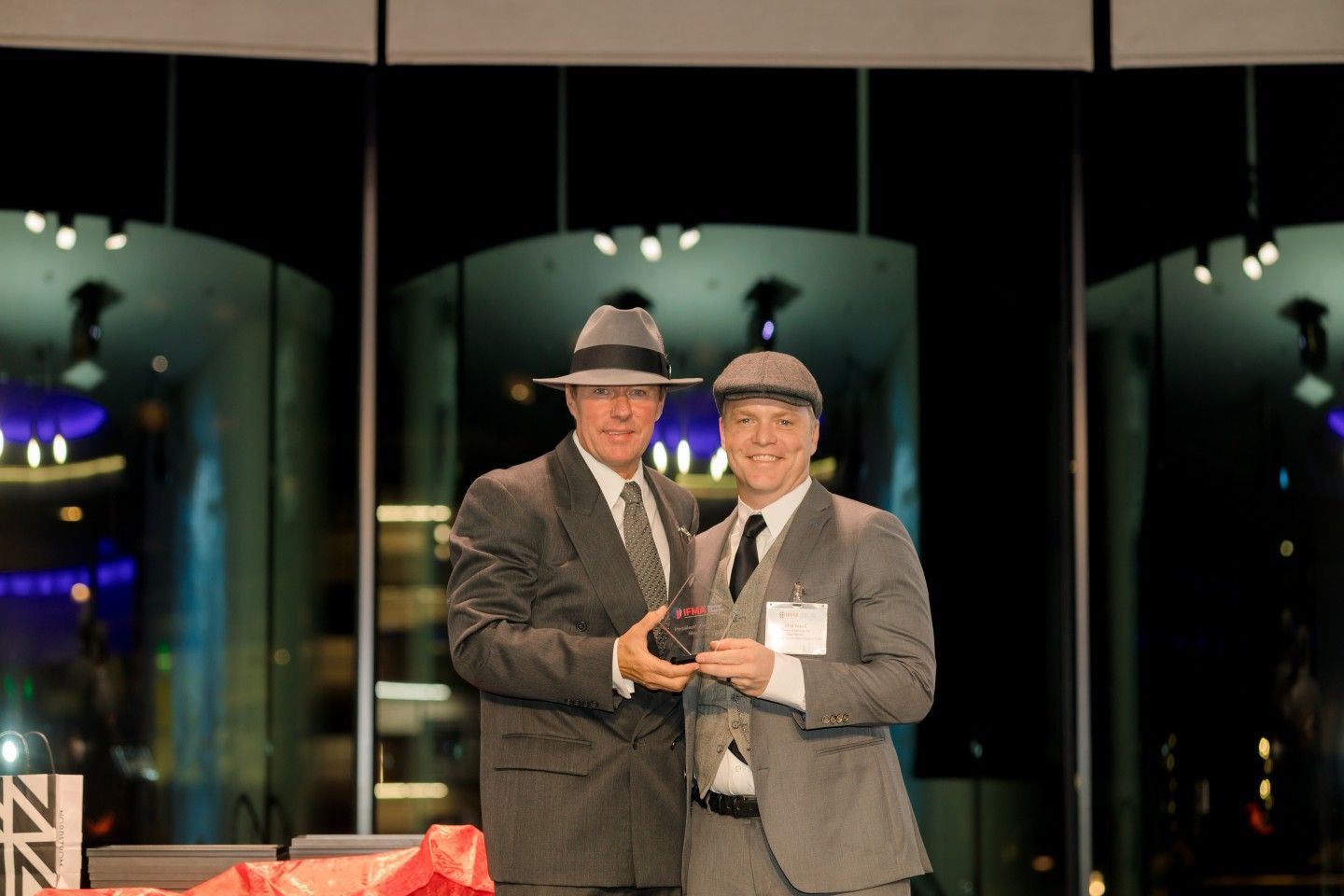 Two men in gray suits and hats, one holding an award, smiling in a modern venue.
