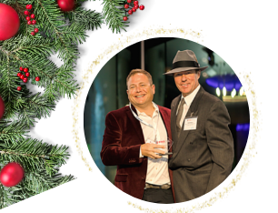 Two men smiling, one in a suit and fedora, the other in a maroon jacket, next to a Christmas tree.