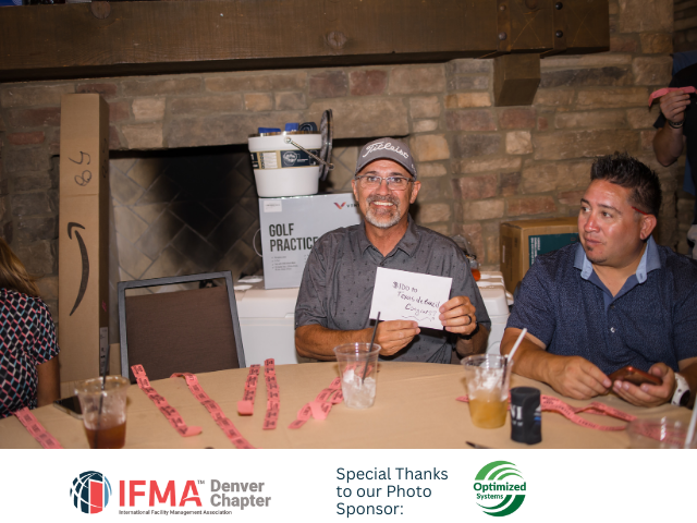 Man holding card smiles, sitting at table with other people and prizes.