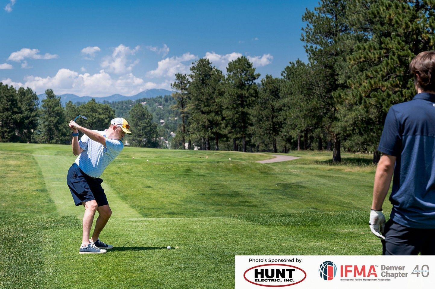 Golfer swinging club on a sunny course, another person watches. Mountains and trees in background.