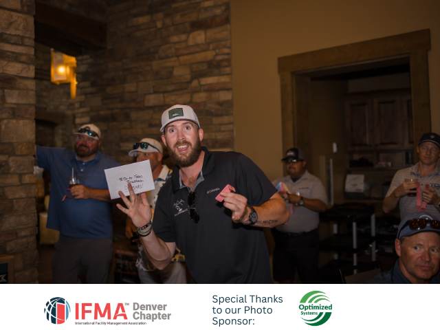 Man in a hat holding a pink item, gesturing excitedly. Group of people in background, indoors.
