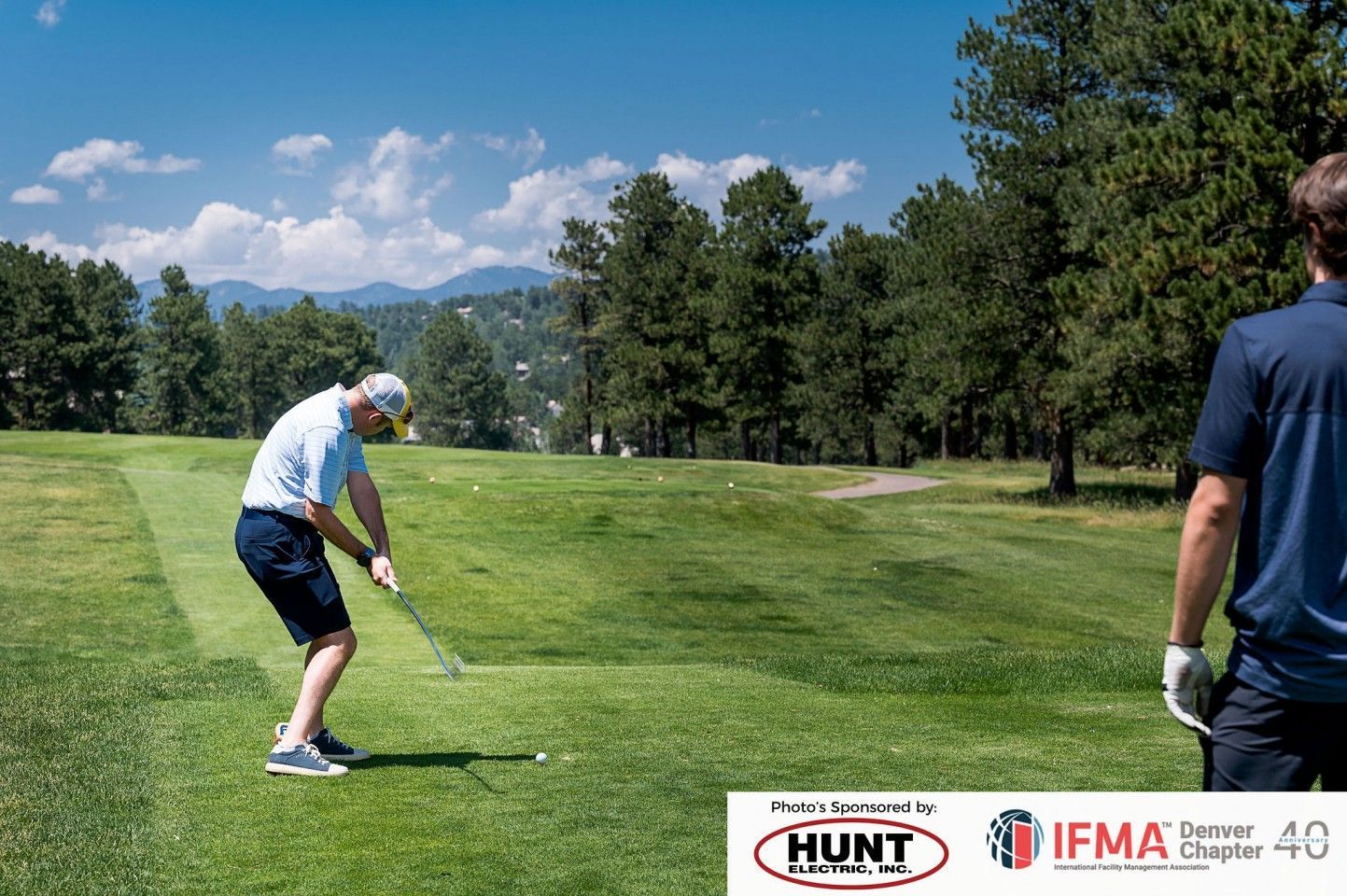 Man swings golf club on green, another watches. Sunny day, trees in background.