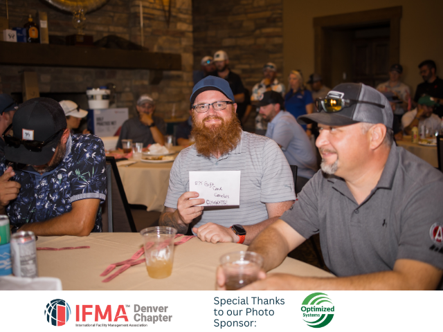 Group seated at a table, man holding card, others smiling. Restaurant setting, Denver Chapter IFMA event.
