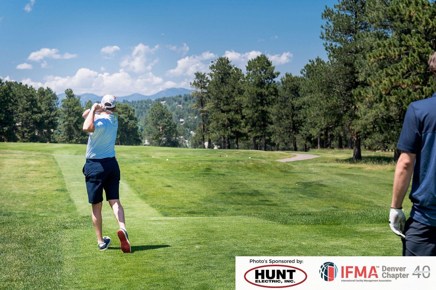 Golfer swinging club on a sunny course, another person watches. Mountains in background.