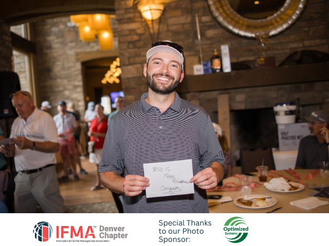Man holding a sign with text, smiling at a golf event. Indoor setting, people in background.