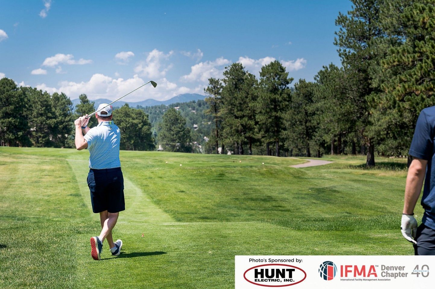 Golfer swinging club on a green course with trees, mountains, and logo in frame.