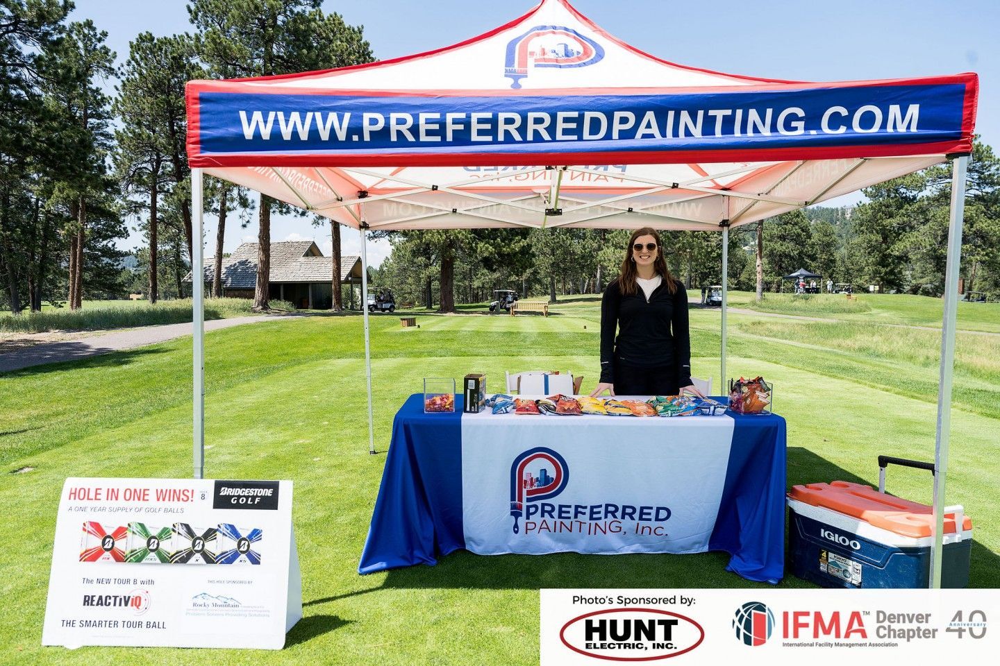 Woman stands at a Preferred Painting booth on a golf course, with website banner and company logo displayed.