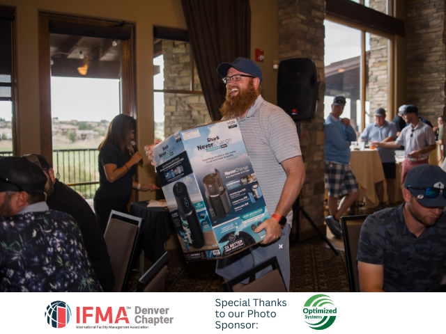Man holding a large box, smiling. Attendees and a speaker present at an event, inside a building with a view.