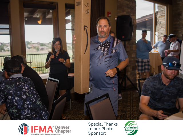 Man carrying a large box at an event; woman with a microphone speaks. People in background.