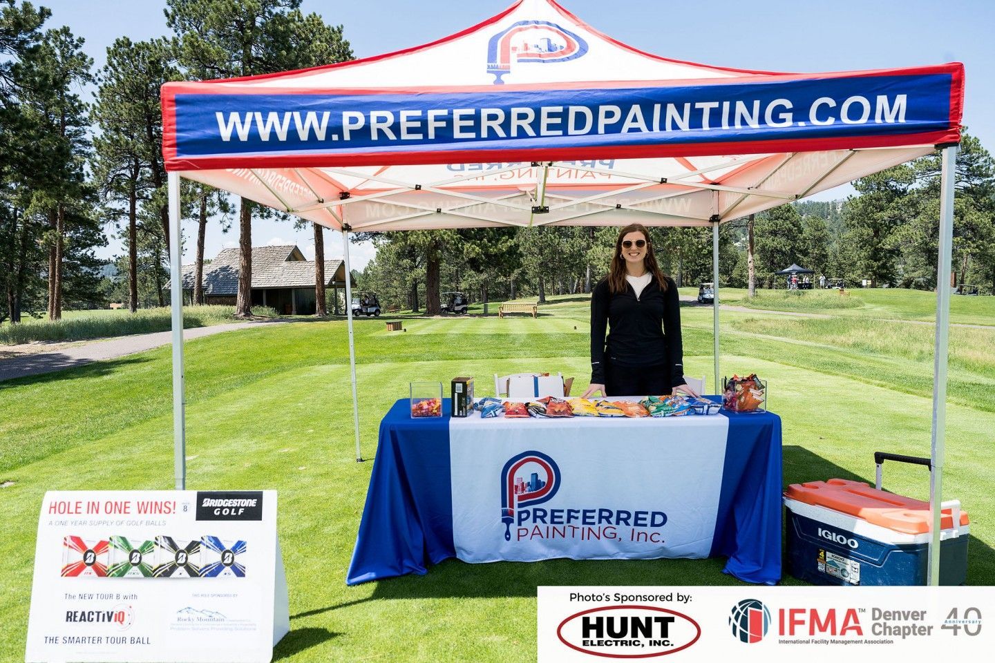 Woman stands at Preferred Painting booth at an outdoor event. Tent, table, and golf course in background.