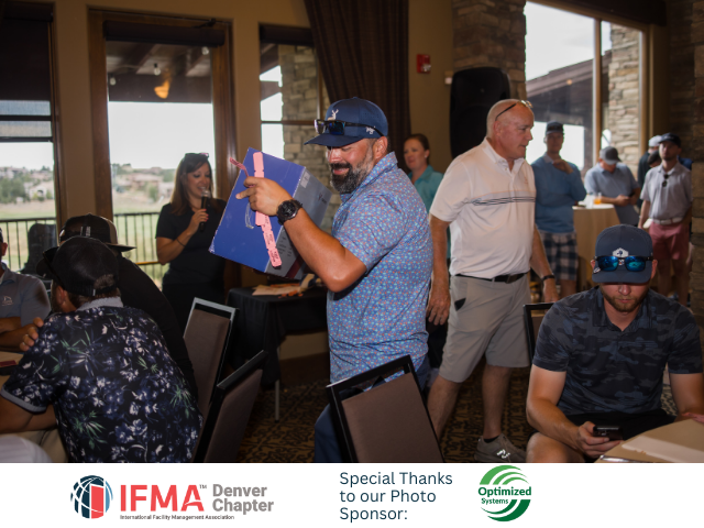 Man in blue cap holds a gift, smiling, indoors. Others watch in a restaurant.