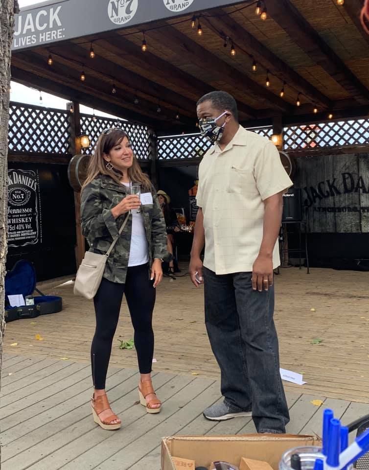 Woman in camo jacket talks with man wearing a mask; outdoor patio with Jack Daniel's branding.