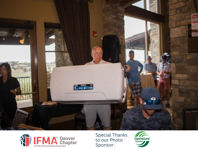 Man holding large white cooler at a golf event, people in background, sunny day.