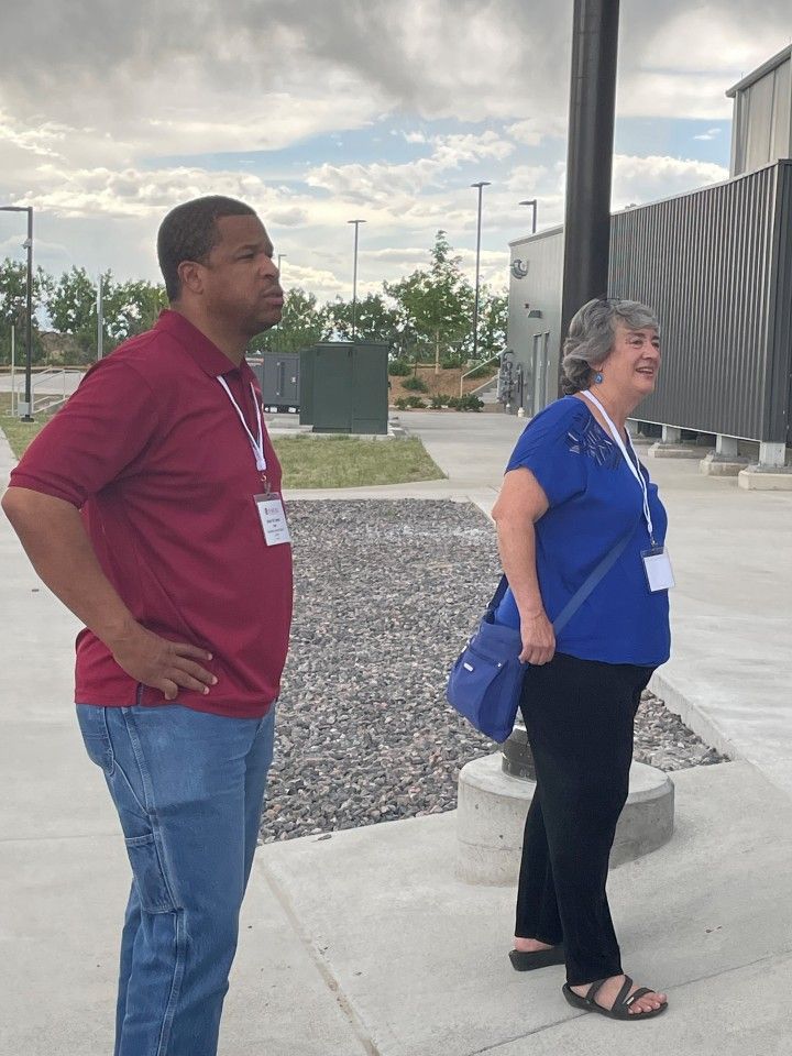Two people standing outside, looking at something. Man in red shirt, jeans. Woman in blue shirt, black pants. Cloudy sky.