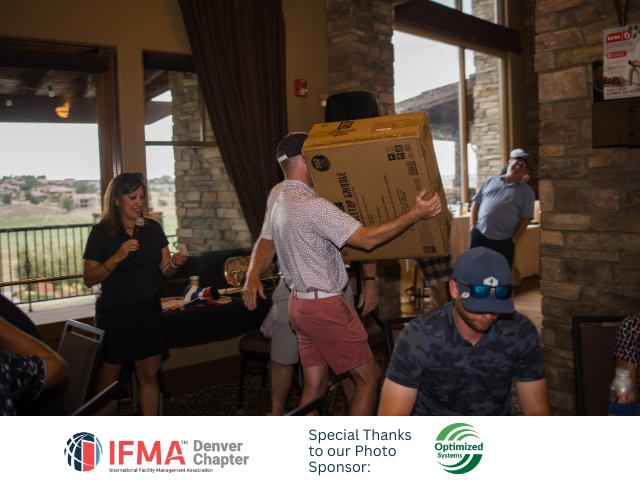 People at an event, one man carrying large box, woman speaking, others watching. Inside a building.