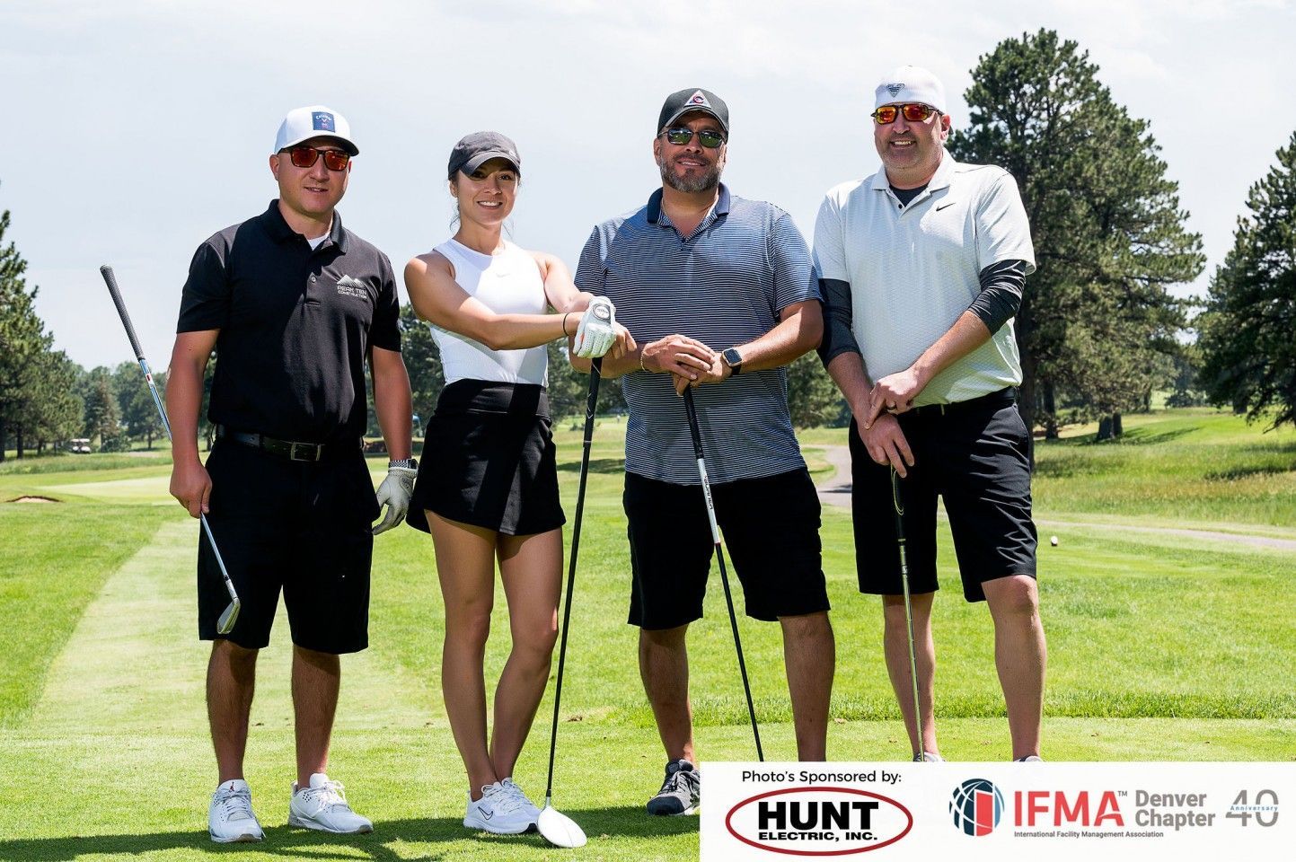 Four people pose on a golf course, holding clubs. Green grass, trees, sunny day.